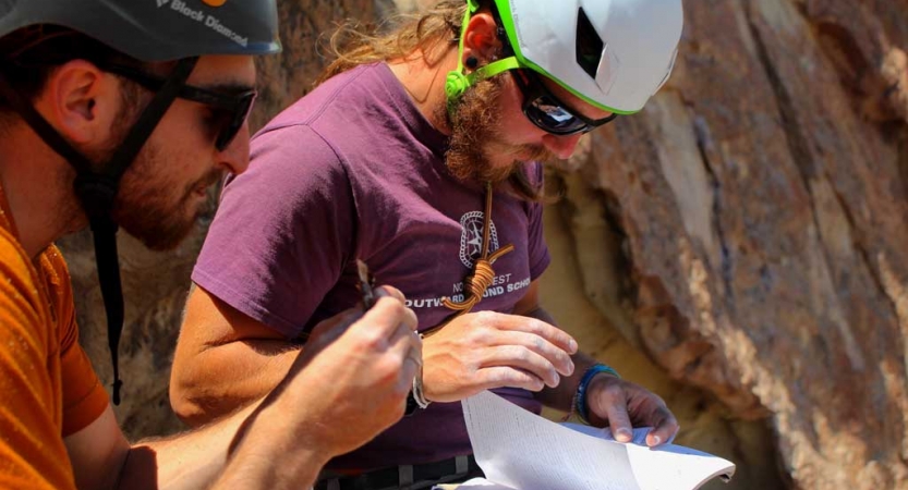 two people wearing helmets examine a book whiles standing next to a rock wall